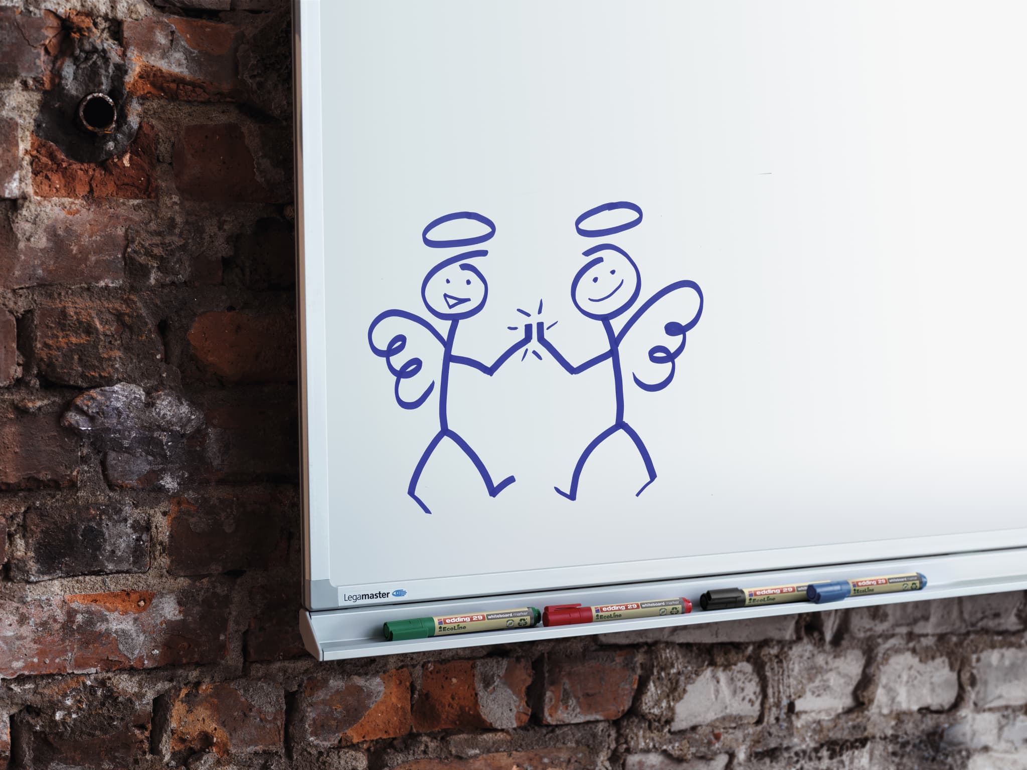 Ein einfaches Gekritzel aus zwei Engelsfiguren mit Heiligenscheinen, Flügeln und lächelnden Gesichtern, gezeichnet mit einem edding 29 EcoLine Whiteboardmarker 4er-Set auf einem Whiteboard vor einem Backsteinmauerhintergrund.