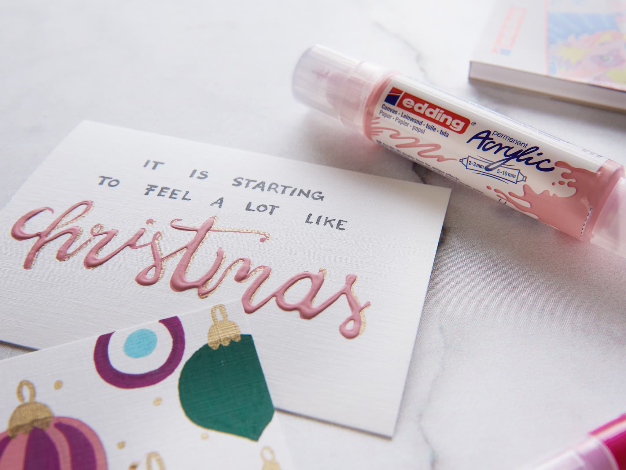 Handschriftliche Festtagsnotiz mit der Aufschrift „Es fängt an, sich ganz nach Weihnachten zu fühlen“ neben einem Metallic-Pink-Marker und Weihnachtskarten auf dem edding 30010 Acryl- und Ölmalblock.