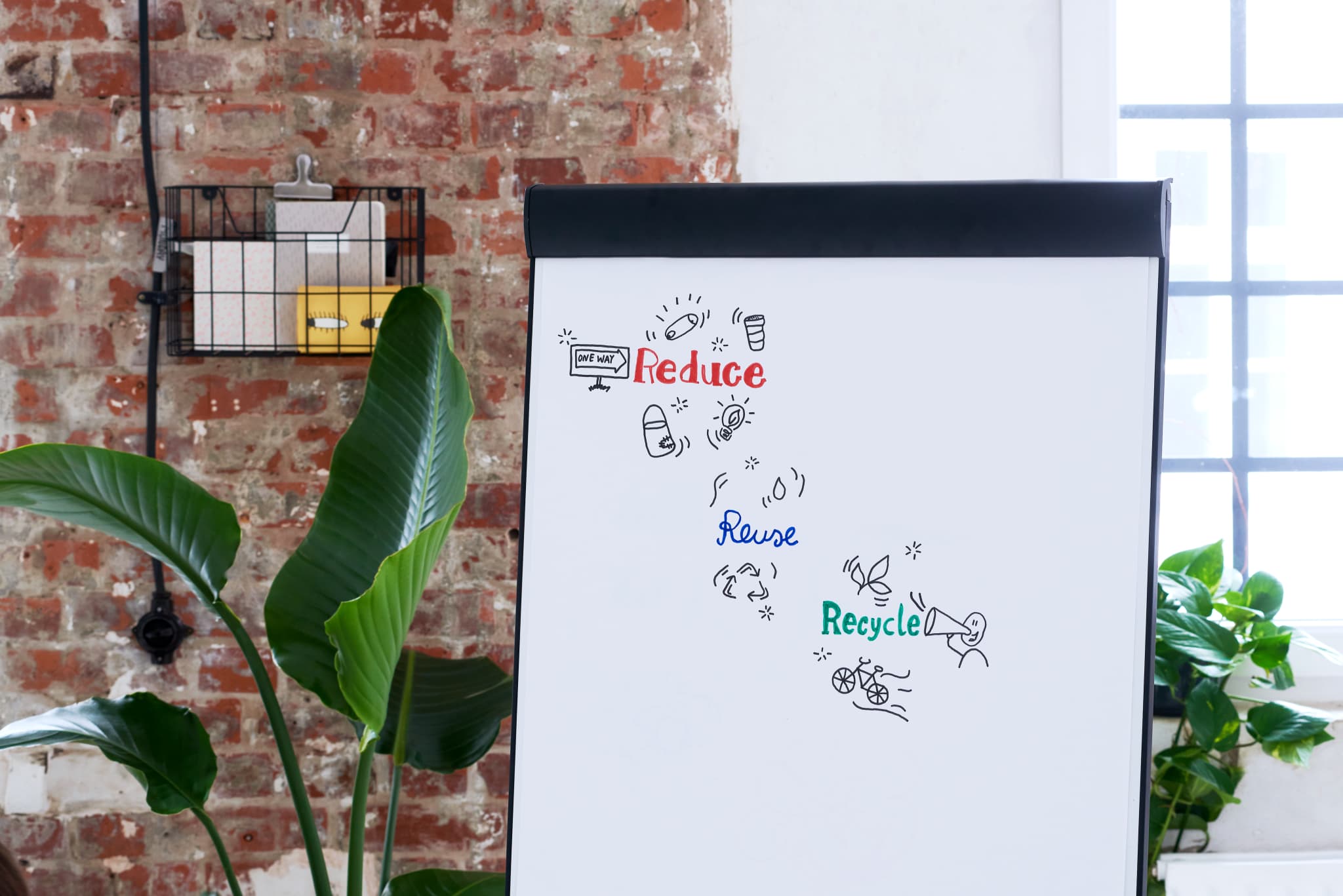 Ein weißes Flipchart mit darauf skizzierten Nachhaltigkeitskonzepten wie „Reduzieren, Wiederverwenden, Recyceln“ mit einem edding 31 EcoLine Flipchartmarker 4er-Set in einem modernen Raum mit Backsteinwänden und einer grünen Pflanze an der Seite.