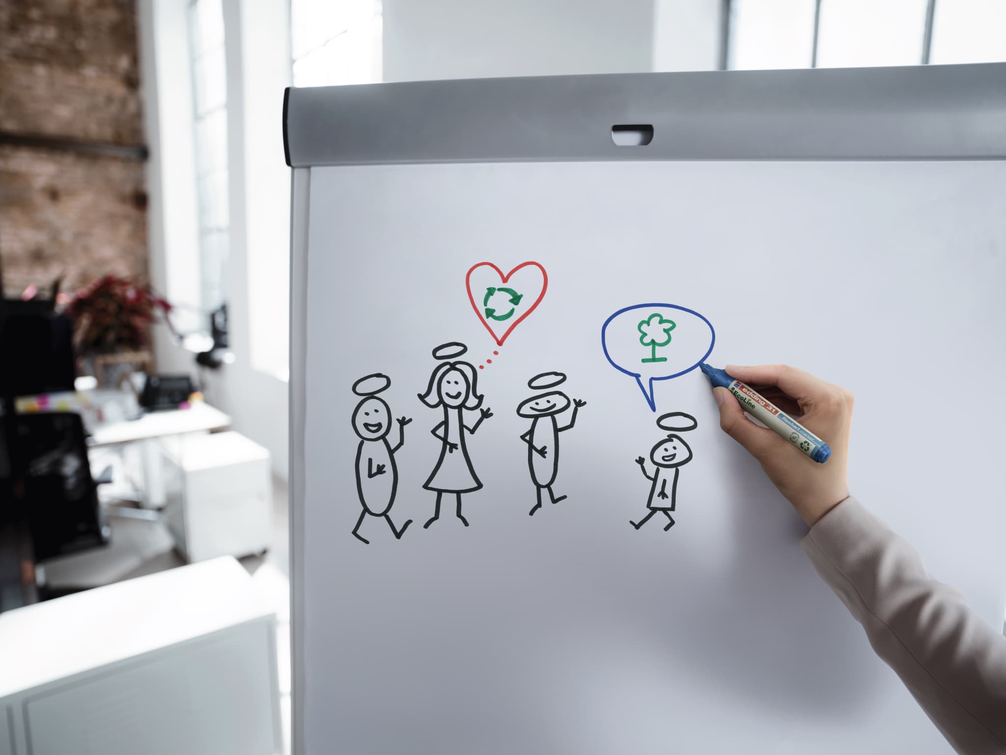 Brainstorming auf einem Whiteboard: Eine Person zeichnet eine Dialogblase mit einem Baumsymbol darin, daneben einfache Strichmännchenzeichnungen einer Gruppe, die über ihrem edding 31 EcoLine Flipchartmarker eine Figur mit einem Herzsymbol enthält.