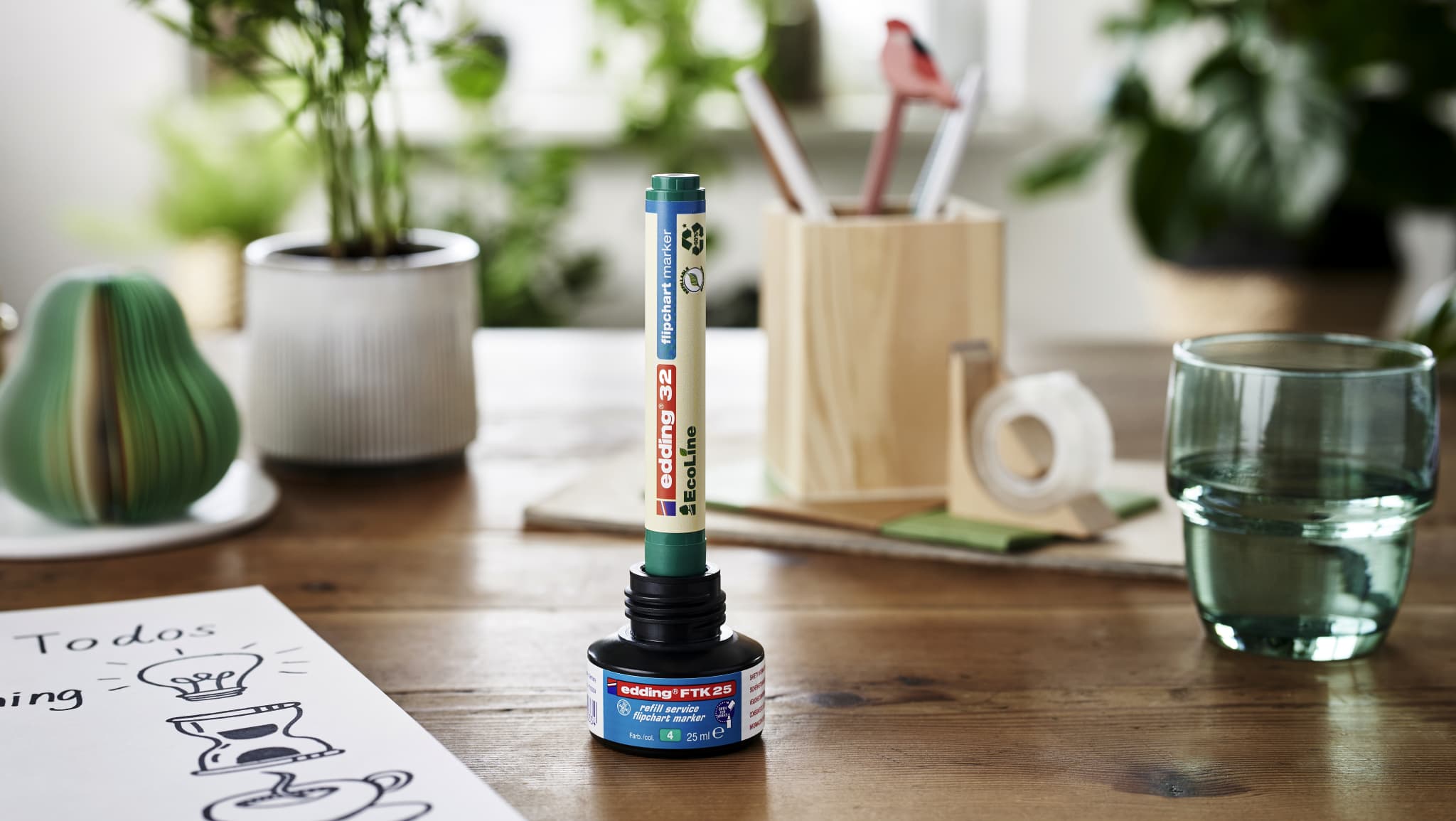Ein gut beleuchteter Arbeitsplatz mit einer Flasche edding EcoLine-Kleber ohne Verschluss und einem aufrecht darauf stehenden Applikator, umgeben von BĂĽromaterialien, einem Glas Wasser und Topfpflanzen.