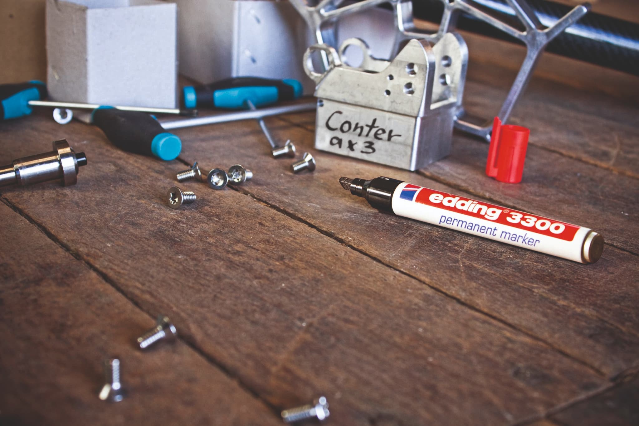 Eine Sammlung verschiedener Werkzeuge und Hardware-Artikel, darunter ein edding 3300 Permanentmarker 4er-Set, verstreut auf einer Holzoberfläche, was auf eine laufende Arbeit oder ein kürzlich abgeschlossenes Projekt schließen lässt.