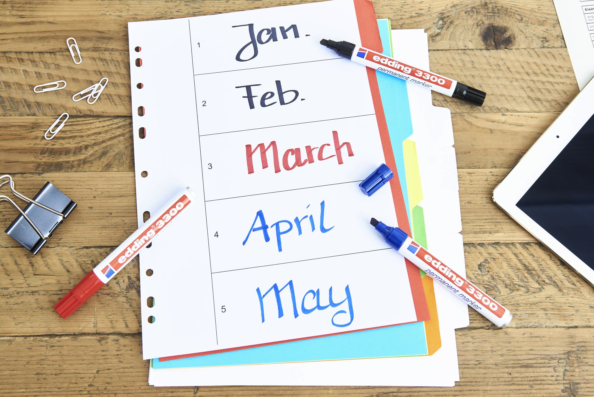 Ein auf einer Holzoberfläche ausgelegter Monatsplaner mit den Monaten Januar bis Mai in verschiedenen Farben, begleitet von einem Satz bunter edding 3300 Permanentmarker-Marker, einem Tablet, Büroklammern und einem.