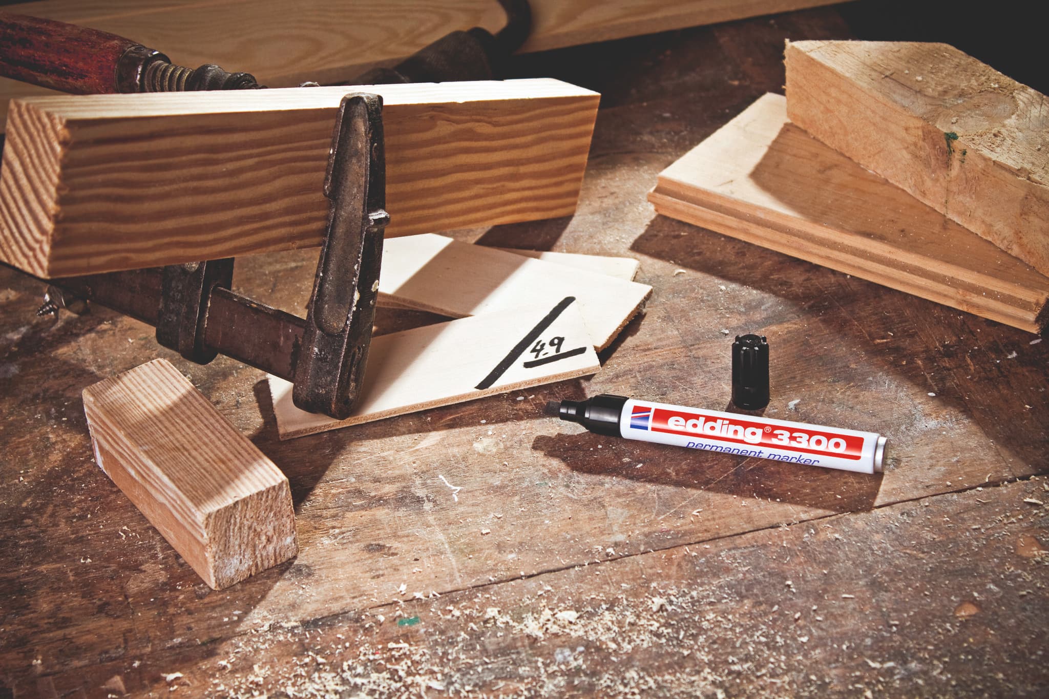 Ein Holzbearbeitungsaufbau mit einer Klemme, die zwei Holzstücke, Späne, ein edding 3300 Permanentmarker 10er-Set zum Markieren von Maßen und ein Blatt Papier mit der Nummer „4,9“ hält.