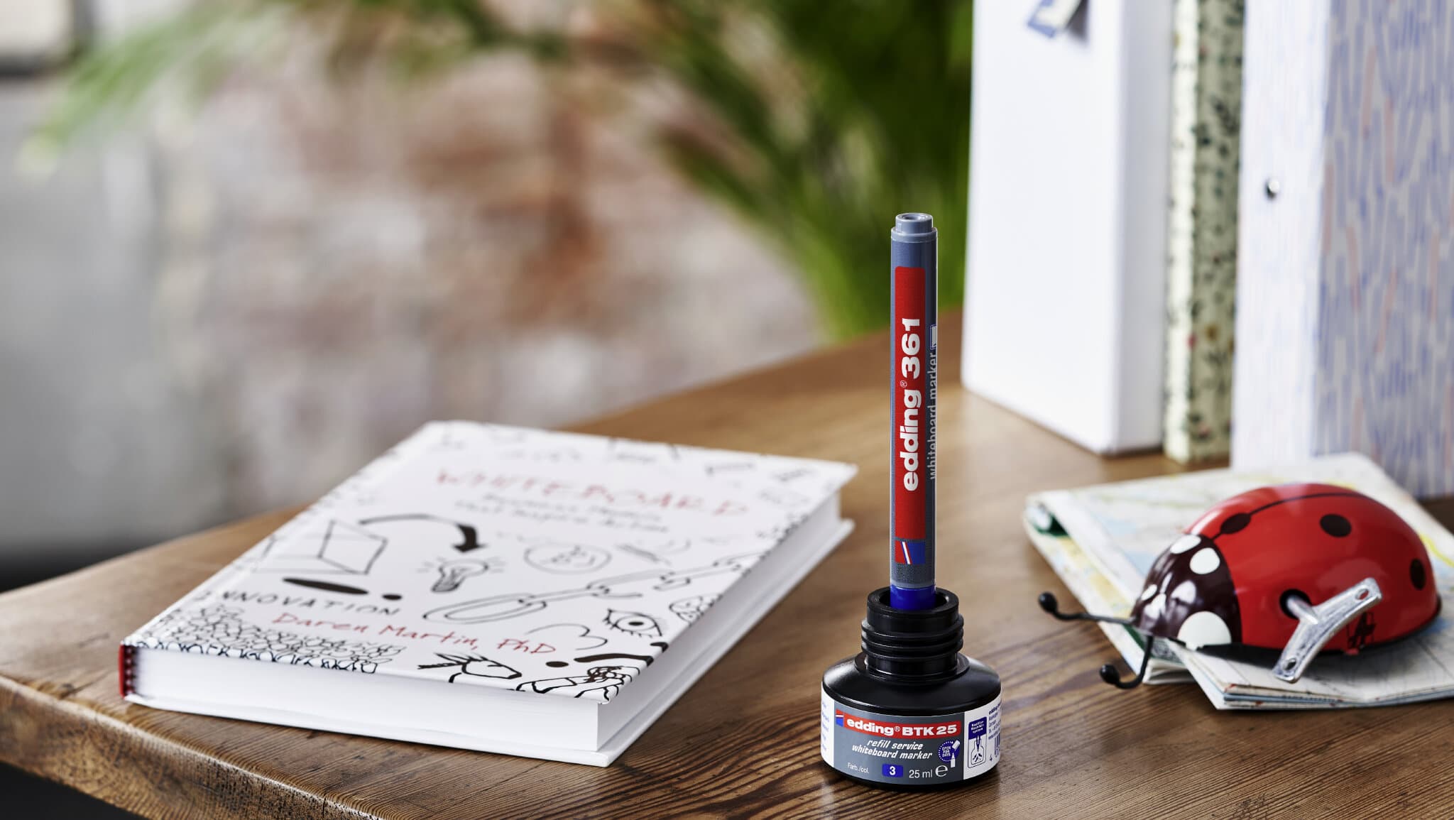 Eine Tube Klebekleber steht aufrecht auf einer Holzoberfläche neben einem aufgeschlagenen Notizbuch mit Kritzeleien und Text im edding 361 Whiteboardmarker 4er-Set, im Hintergrund teilweise sichtbar ein Fahrradhelm.