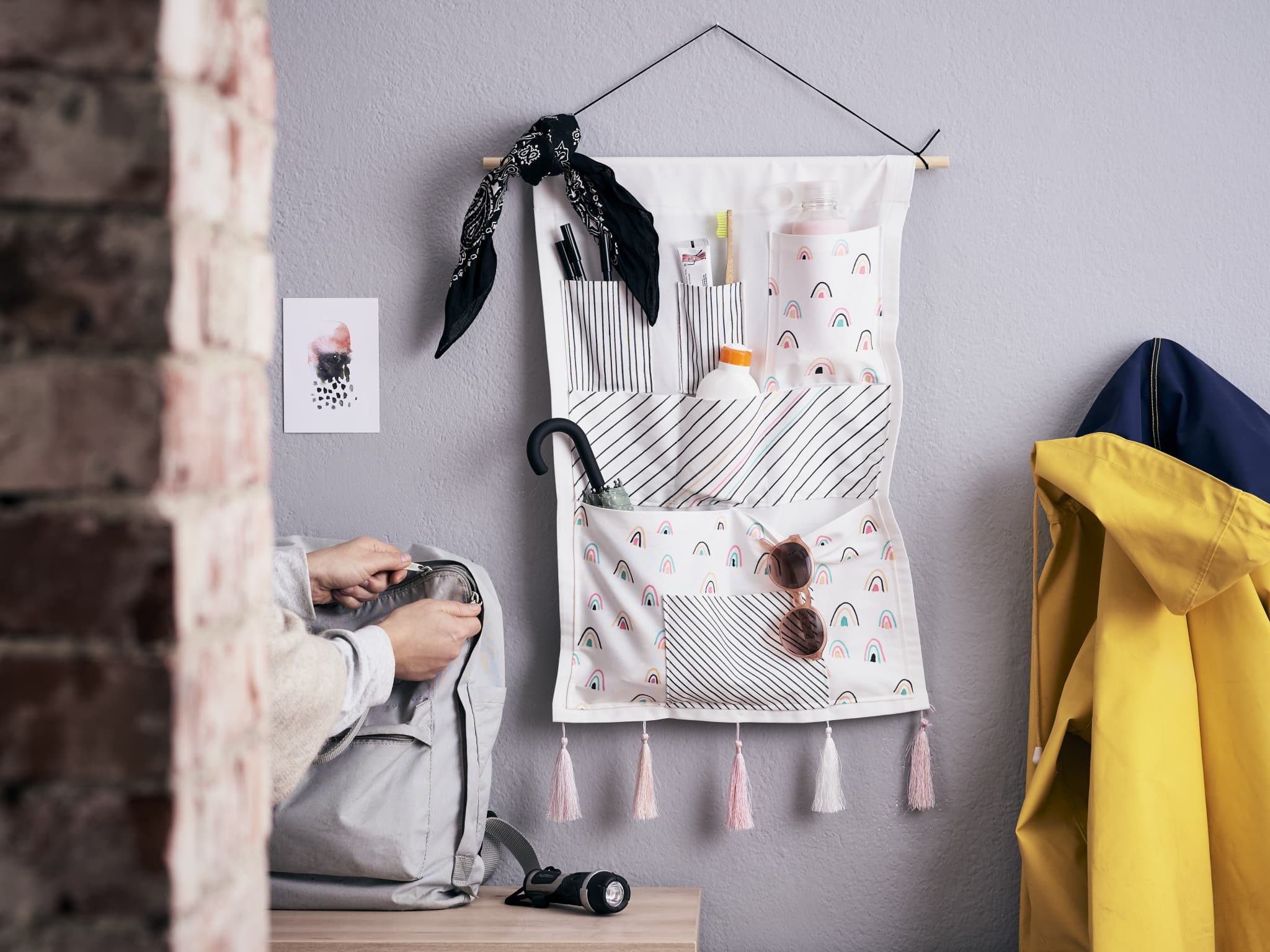 Eine gemütliche Ecke mit einem stilvollen Wandorganizer mit Taschen, einem gelben Mantel, einer sitzenden Person mit einer Kamera und einem schwarzen Regenschirm, der neben Dekorationsgegenständen mit einzigartigen edding 4600 Textilstift-Designs hängt.