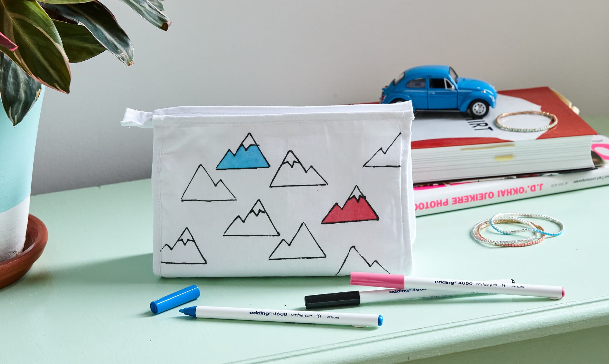 Ein weißes Federmäppchen mit einem edding 4600 Textilstift-Muster liegt auf einem Stapel Bücher neben bunten Markern auf einem mintgrünen Schreibtisch, während ein blaues Miniaturauto und eine Pflanze für einen Hauch von Laune sorgen.