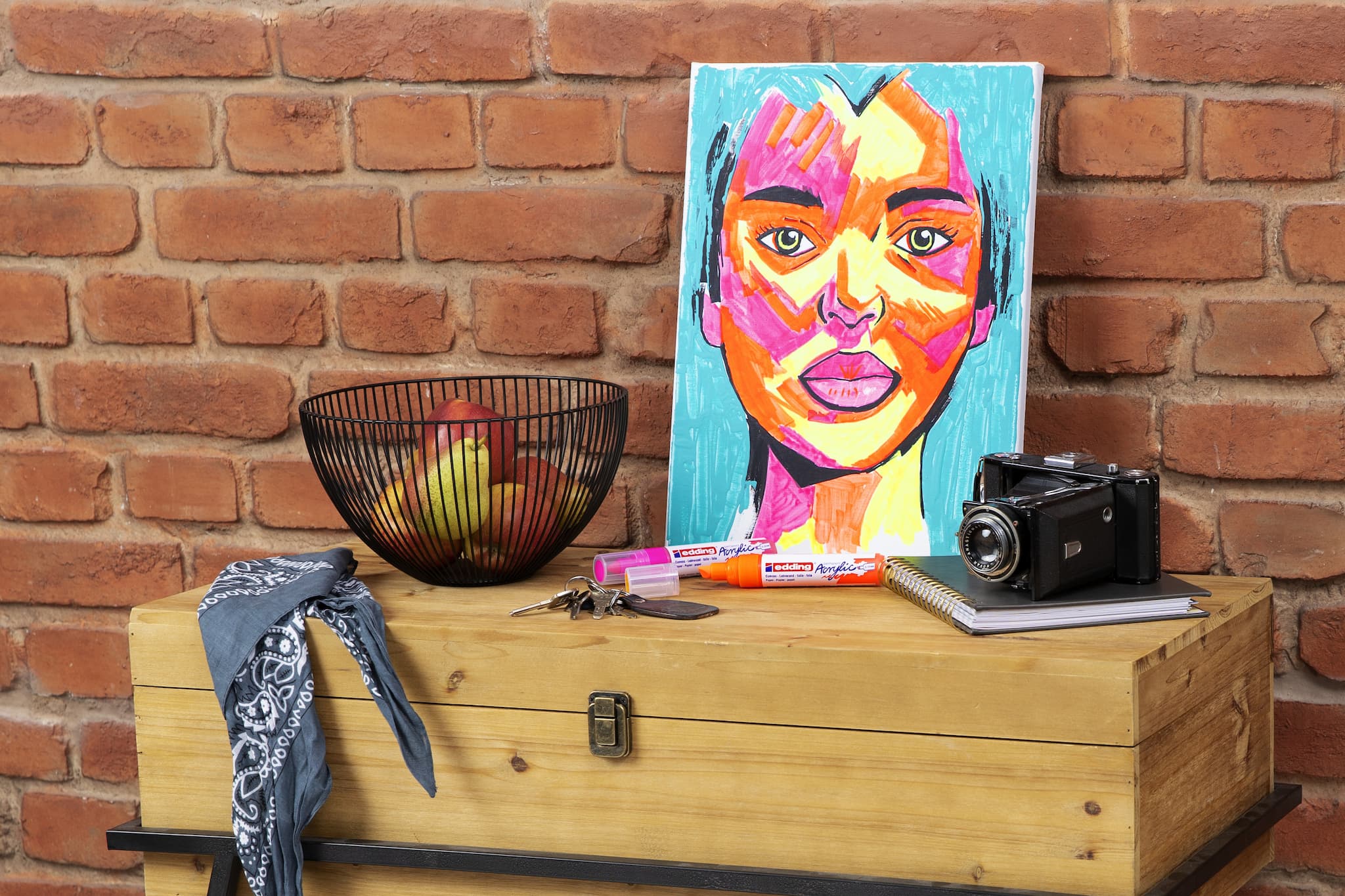 Ein farbenfrohes abstraktes Porträtgemälde, das mit dem edding 5000 Acrylmarker breit 5er-Set erstellt wurde, ruht auf einer Holzkiste an einer Ziegelwand, begleitet von einer Auswahl an Gegenständen, darunter eine Kamera, eine Obstschale und Malutensilien.