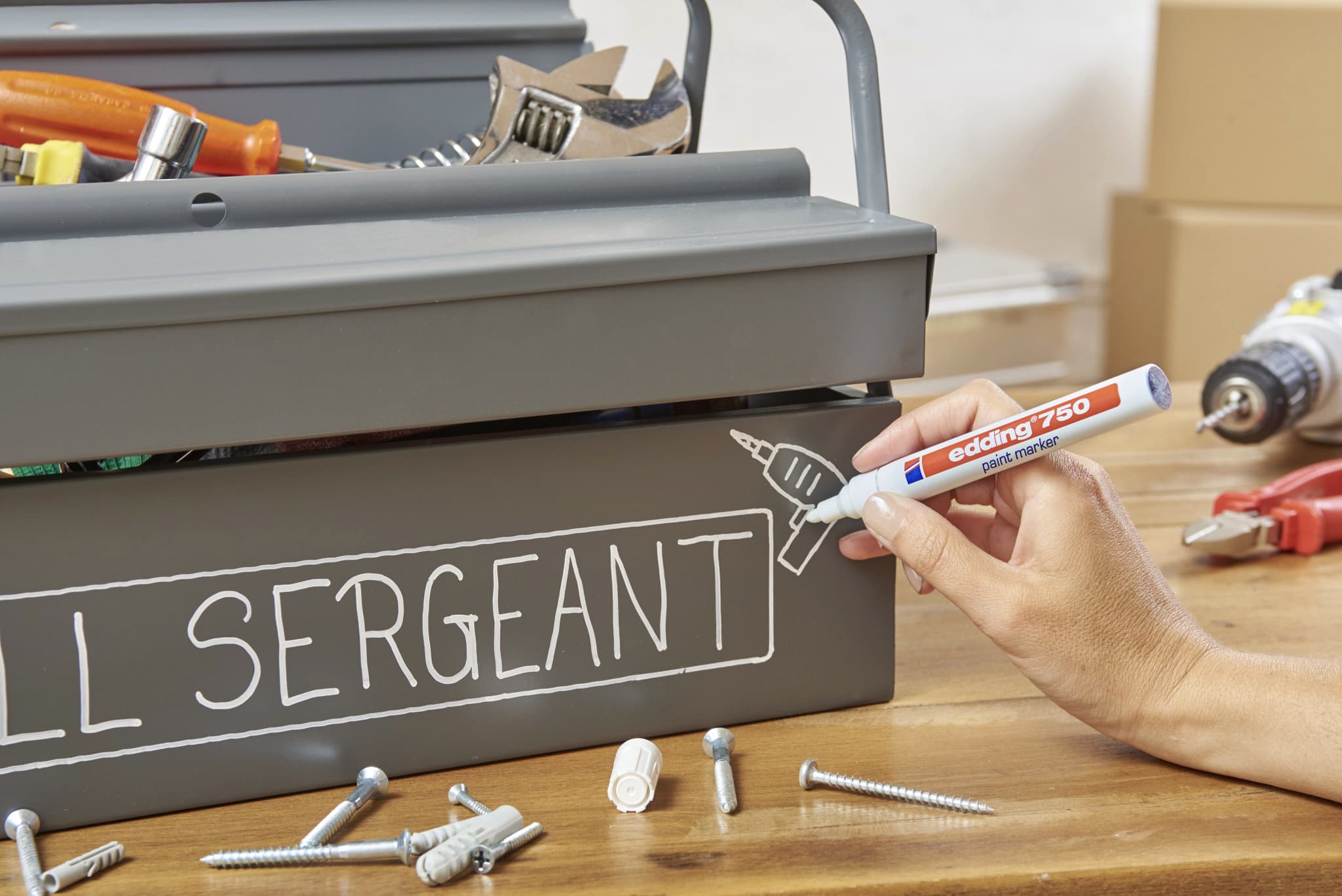Eine Hand beschriftet mit dem edding 750 Lackmarker 2er-Set von edding einen grauen Werkzeugkasten mit „SERGEANT“ und einer Bohrerzeichnung, während daneben auf der Holzfläche verstreut diverse Werkzeuge und Schrauben liegen.