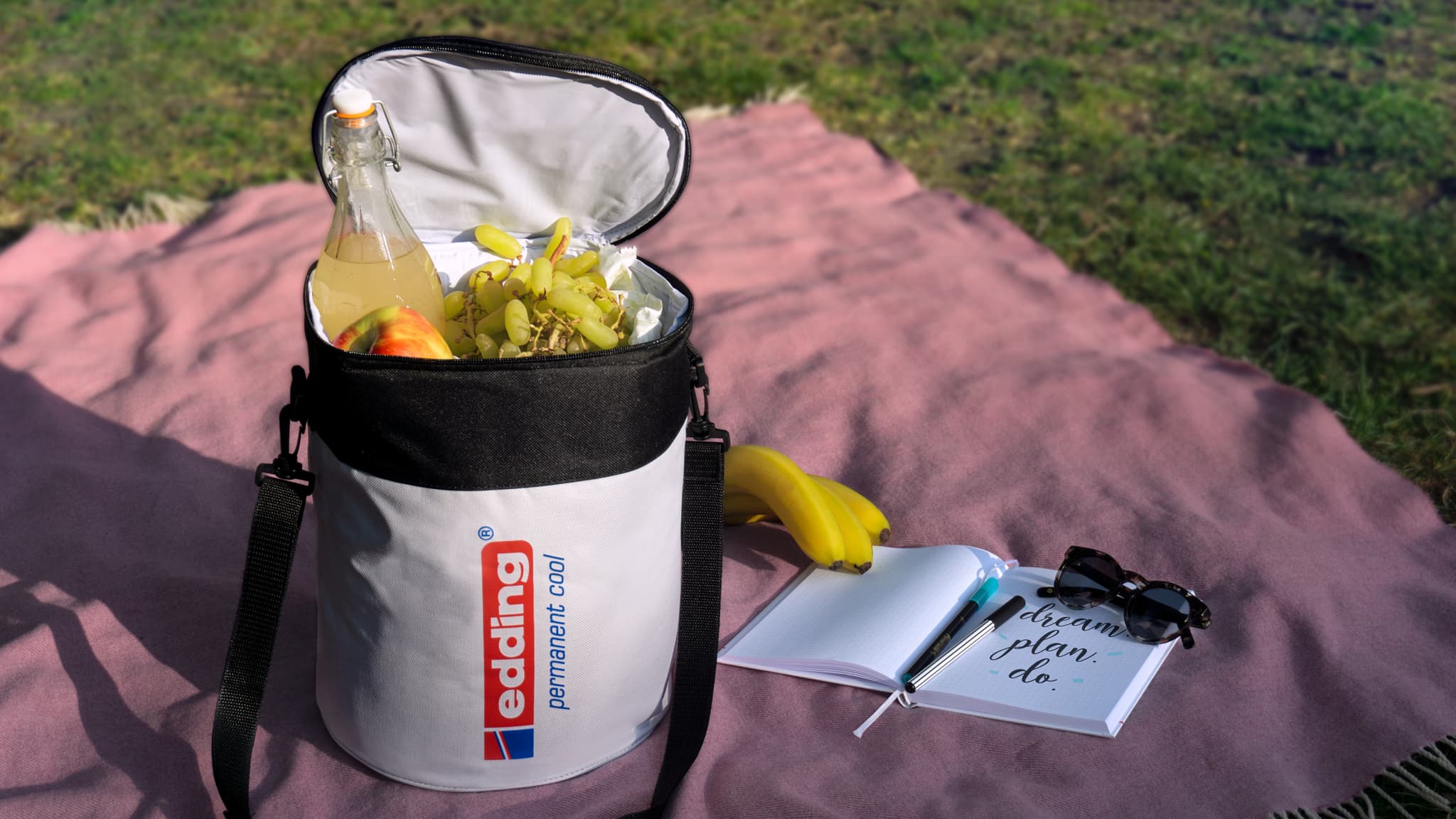 Eine zylindrische weiße edding Kühlbox mit einem schwarzen Oberteil, gefüllt mit rotkappigen Flaschen auf einem schwarzen H.