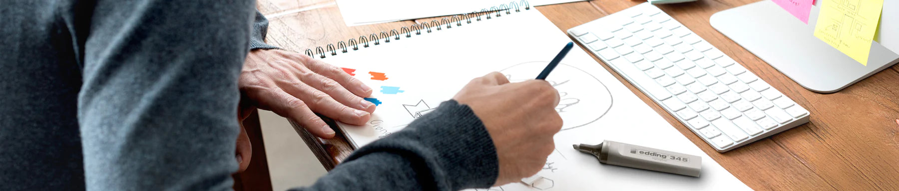 Eine Person skizziert ein Designkonzept auf einem Notizblock neben einer Tastatur, wobei verschiedene Designwerkzeuge und Haftnotizen in Reichweite sind.