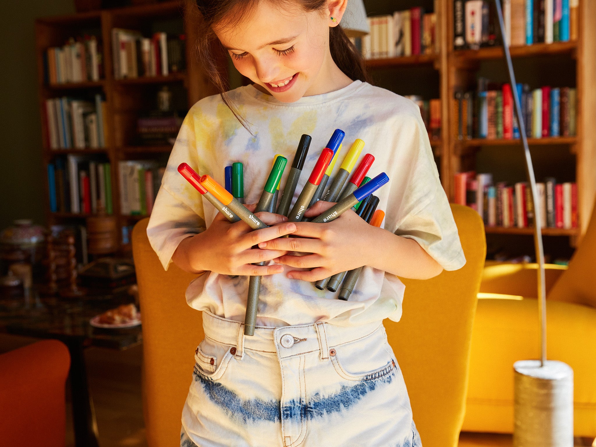 Ein junges Mädchen mit strahlendem Lächeln hält eine bunte Sammlung von Markern an die Brust und steht in einem gemütlichen Raum voller Bücher.