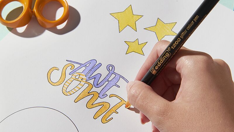 Die Hand einer Person, die mit einem schwarzen Feinpunktmarker Details zu einem farbenfrohen, handgeschriebenen Kunstwerk mit der Aufschrift „awesome“ hinzufügt, umgeben von gezeichneten gelben Sternen, daneben liegt eine orangefarbene Schere.