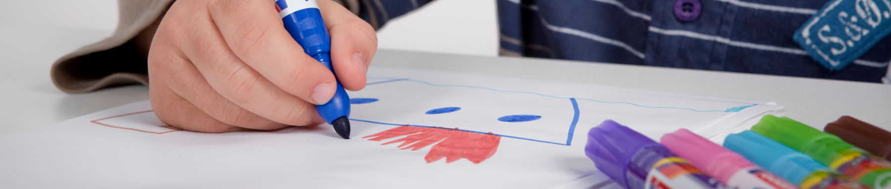 Die Hand eines Kindes, die einen blauen Stift hält, zeichnet ein lächelndes Gesicht auf ein Blatt Papier, umgeben von einer Reihe bunter Stifte.