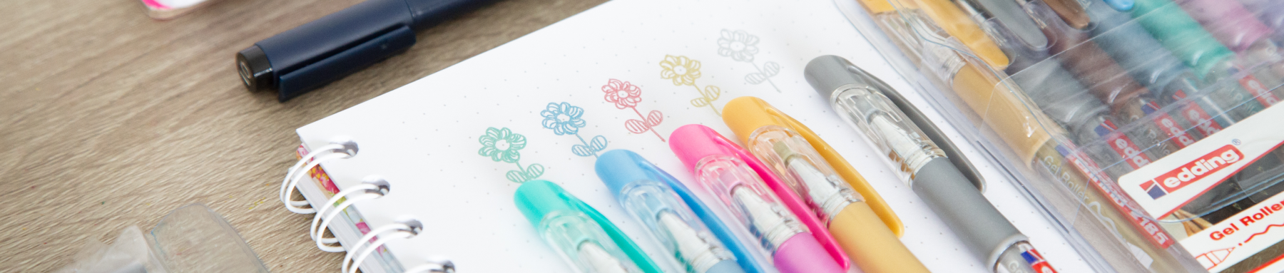 Eine Reihe bunter Stifte liegt neben einem Notizbuch mit gekritzelten Blumen und signalisiert einen Moment der Kreativität oder Planung.