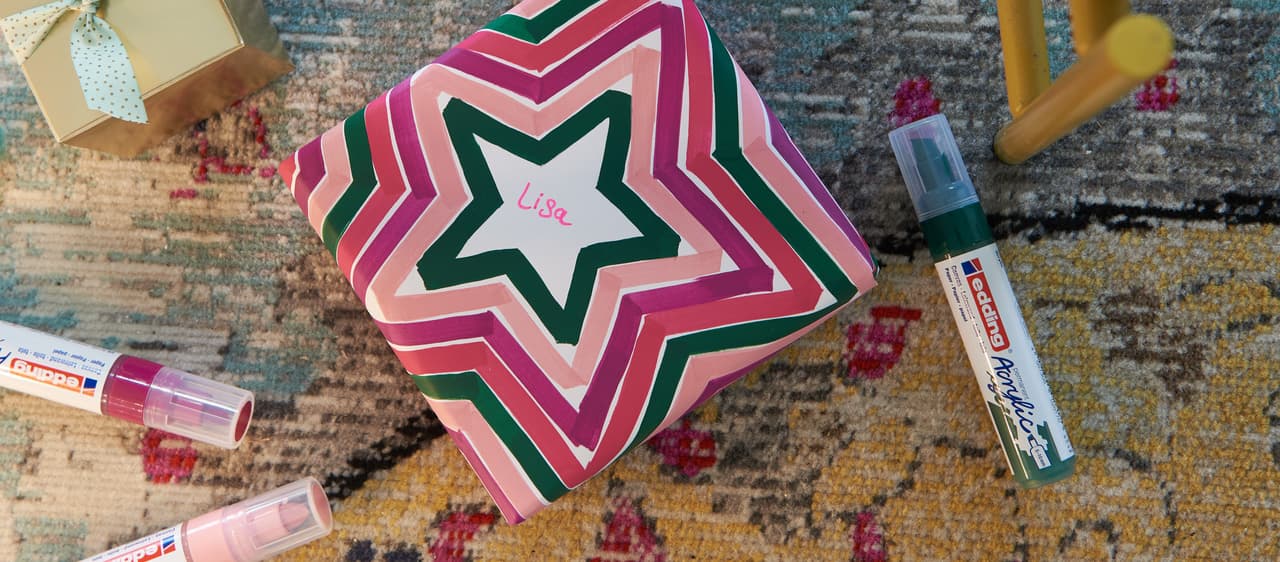 Eine farbenfroh gemusterte Geschenkbox mit dem Namen „lisa“, umgeben von bunten Markern und Lippenbalsam auf einem strukturierten Teppich.