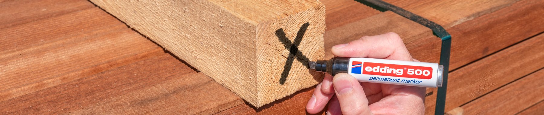 Eine Person markiert mit einem edding 500 Permanentmarker ein schwarzes „x“ auf einem Holzbrett.