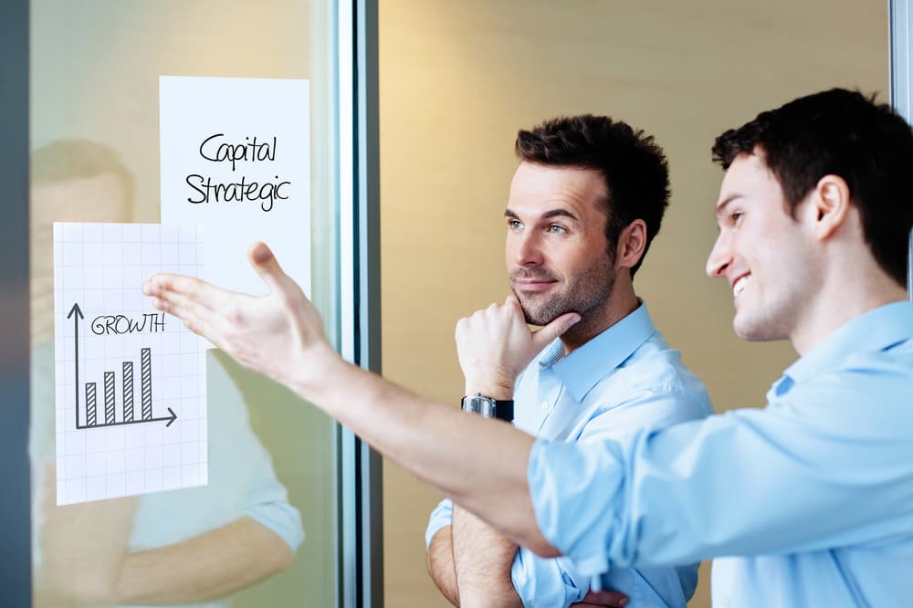 Zwei Männer in blauen Hemden diskutieren in einem Büro über Geschäftsstrategien. Einer zeigt auf ein Whiteboard mit Legamaster Magic-Chart-Notizen (Whiteboard Folie A4), auf denen ein Balkendiagramm mit Wachstumsdaten zu sehen ist, und einer weiteren Notiz mit der Aufschrift „Capital Strategic“. Beide sind engagiert und nachdenklich.