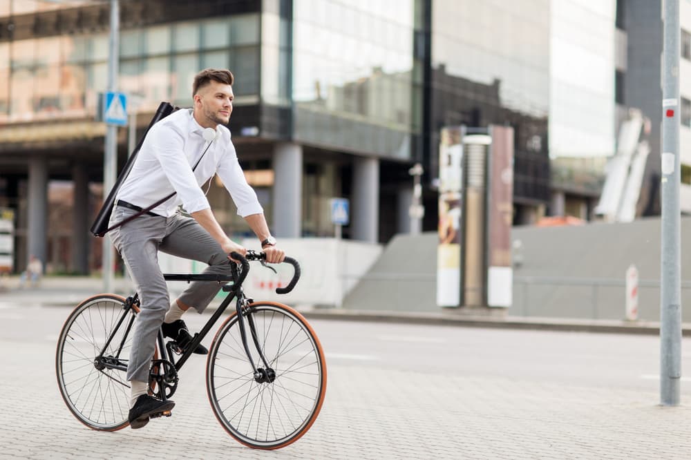 Ein Mann mit weißem Hemd und hellgrauer Hose fährt mit dem Fahrrad durch eine Stadtstraße. Er trägt einen schwarzen Rucksack mit verstellbarem Schultergurt und hat Kopfhörer um den Hals. Im Hintergrund sind Bürogebäude und städtische Infrastruktur zu sehen. Außerdem ist zu sehen, dass er den Legamaster Magic-Chart Transportköcher von Legamaster trägt.