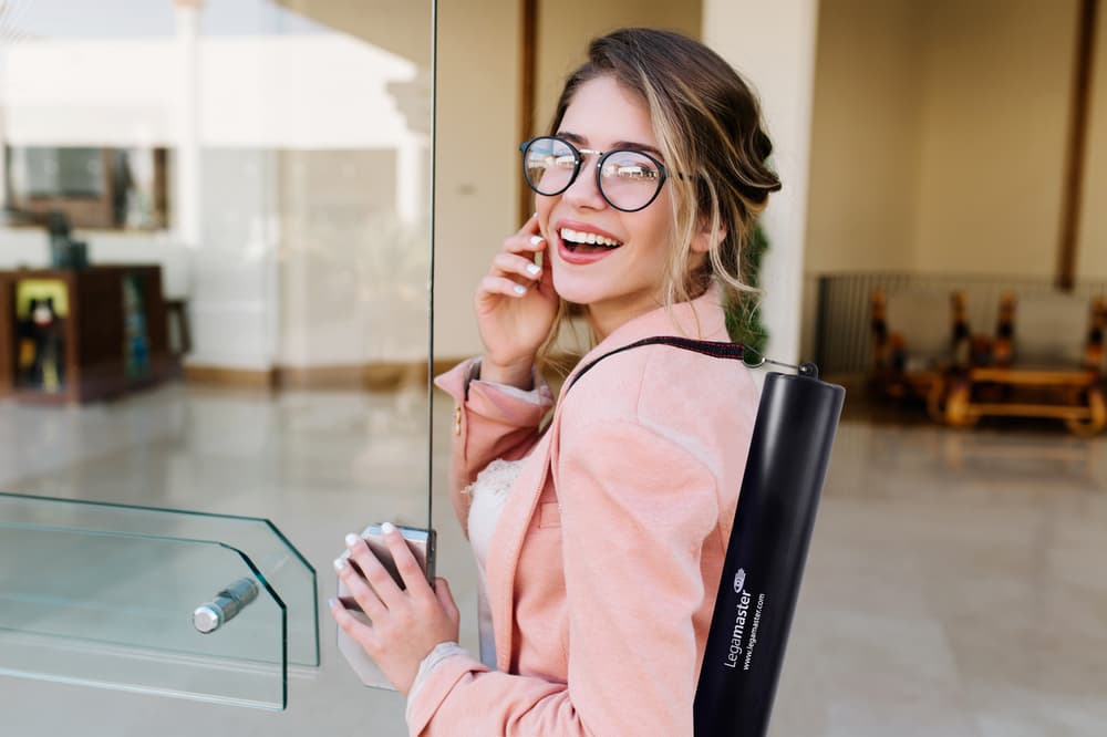 Eine lächelnde Person mit langen Haaren und Brille, die eine rosa Jacke trägt, telefoniert mit einem Mobiltelefon. Sie trägt einen Legamaster Magic-Chart Transportköcher mit einem verstellbaren Gurt auf dem Rücken und steht in einem modernen Gebäude mit Glastüren.