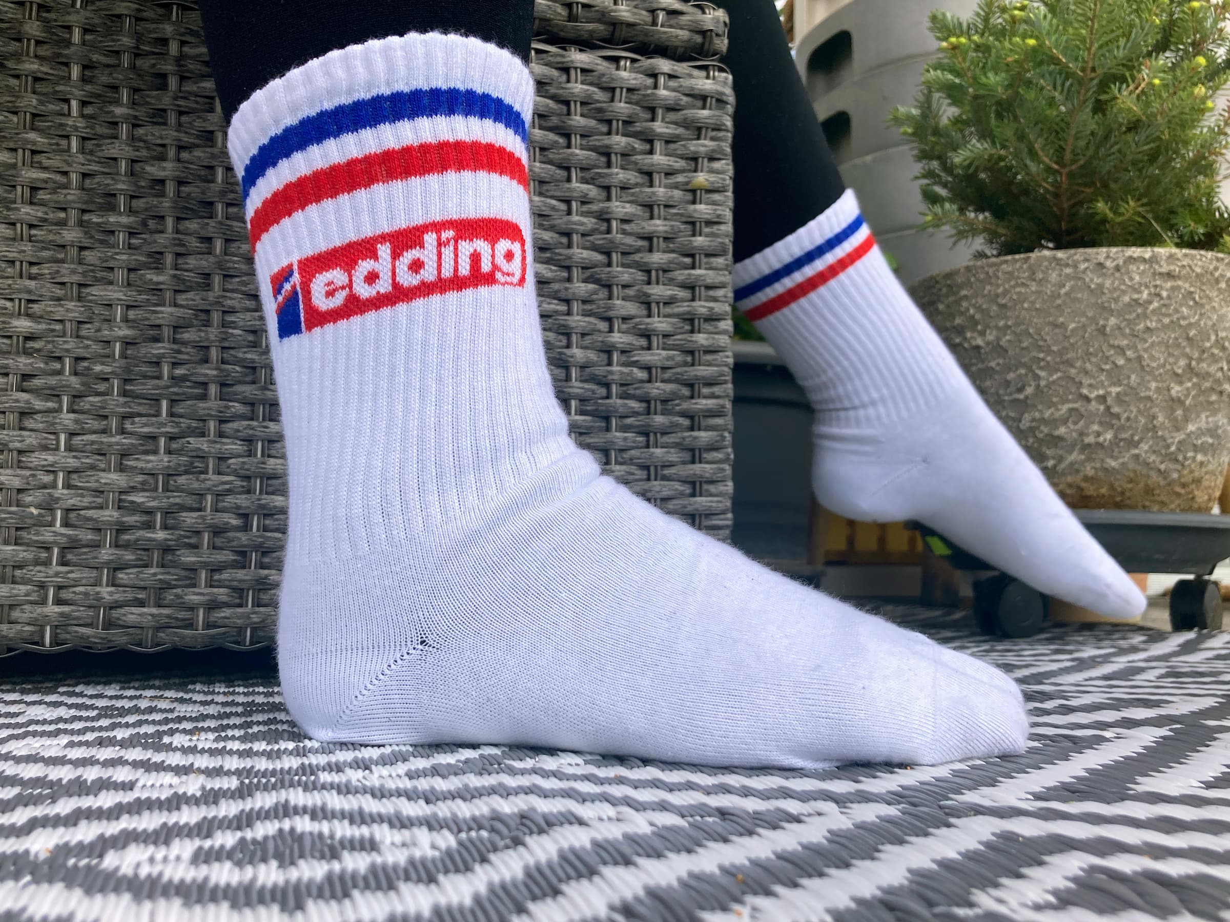 Eine Person trägt Unisex-Tennissocken von edding mit roten, weißen und blauen Streifen und dem Markennamen „edding“ in Rot auf den Seiten. Die Füße der Person liegen auf einem Korbstuhl oder Ottomanen, im Hintergrund ein gemusterter Teppich und eine Topfpflanze. Diese Socken werden in Deutschland hergestellt.