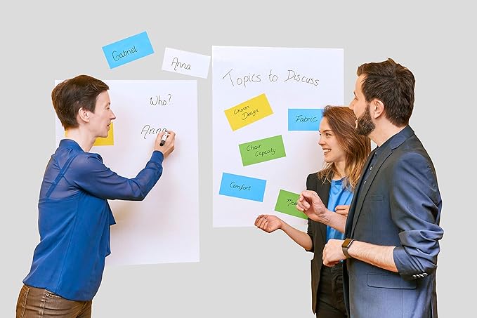 Drei Personen diskutieren an zwei Whiteboards. Eine Frau links schreibt Namen auf ein Whiteboard, während zwei andere, ein Mann und eine Frau, an einem anderen mit der Aufschrift „Themen zur Diskussion“ stehen. Das Board ist mit Legamaster Magic-Chart-Notizen 10x20 cm sortiert von Legamaster bedeckt, mit farbenfrohen statischen Haftnotizen.