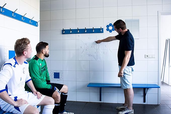 Ein Trainer im schwarzen Hemd steht in der Umkleidekabine und zeigt auf ein Strategiediagramm, das auf einer Legamaster Magic-Chart Whiteboard-Folie gezeichnet ist. Zwei Fußballspieler in Uniformen, einer in einem weißen und einer in einem grünen Trikot, sitzen auf einer Bank und betrachten das Diagramm aufmerksam.