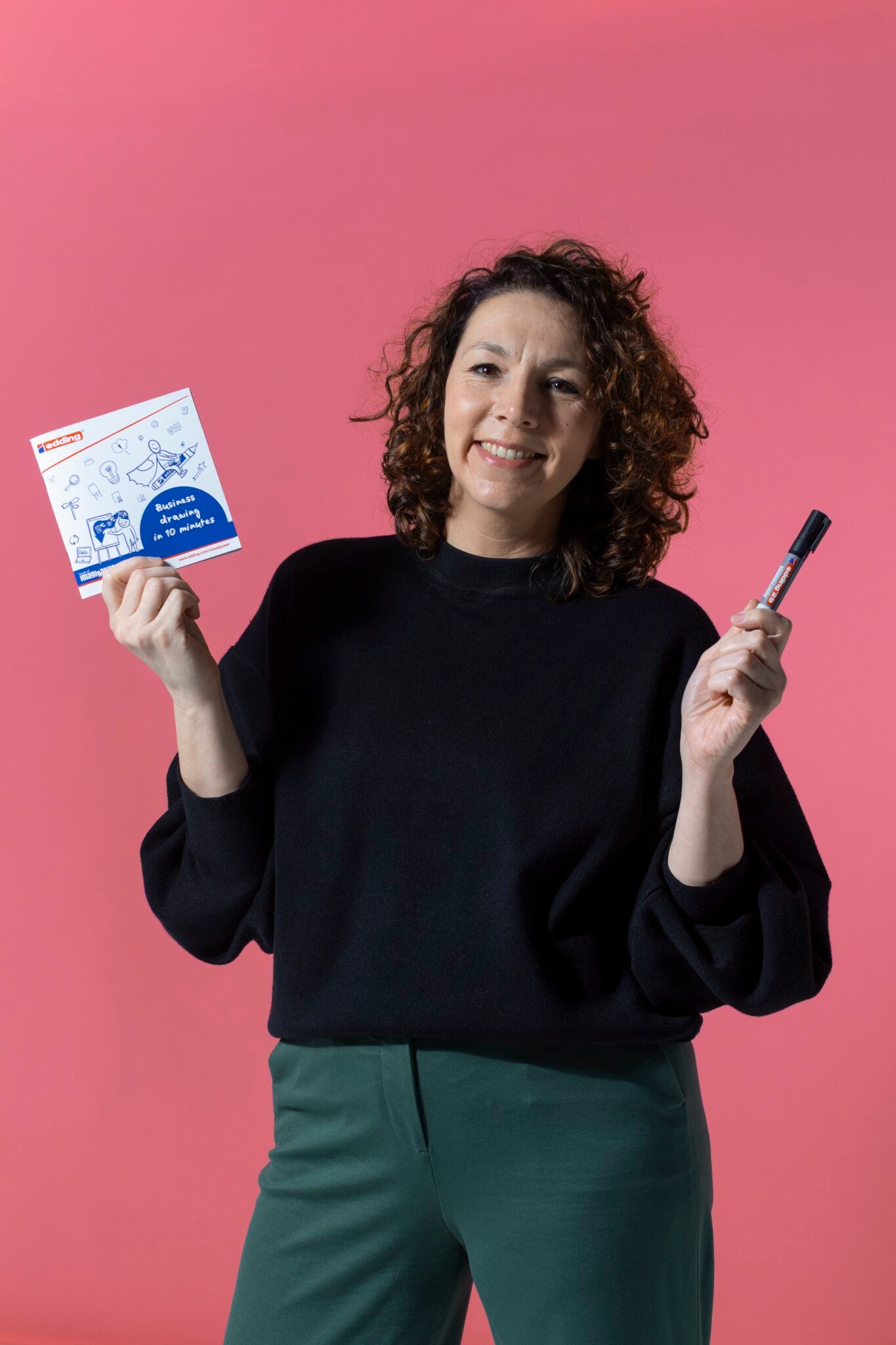 Eine lächelnde Frau mit lockigem Haar steht vor einem rosa Hintergrund und hält die PLAYROOM ESSENTIALS Kreativbox Visualisierung von Playroom. Sie trägt ein schwarzes Oberteil und eine grüne Hose.
