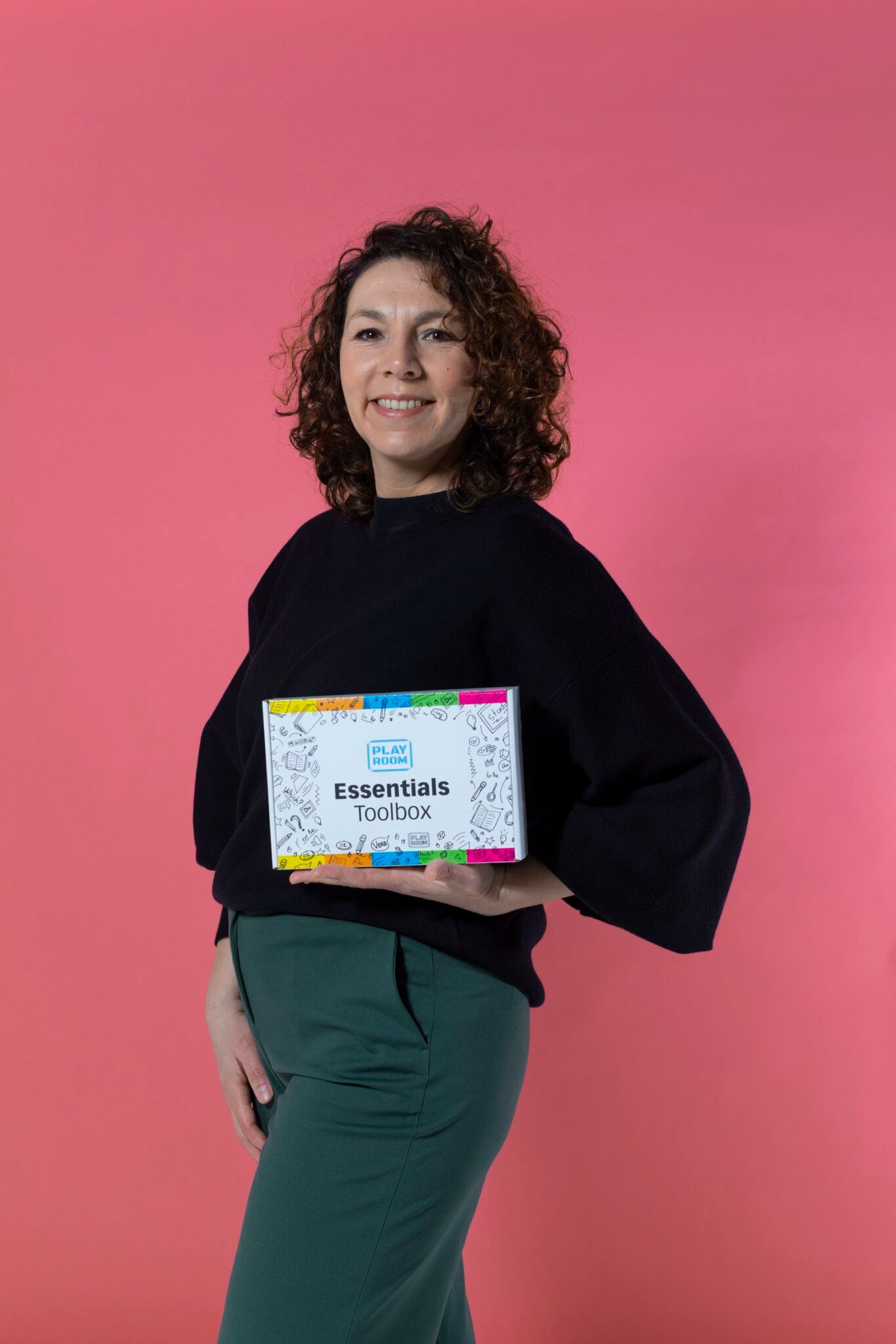 Eine Frau mit lockigem Haar steht vor einem rosafarbenen Hintergrund, lächelt und hält eine Box mit der Aufschrift "PLAYROOM ESSENTIALS Kreativbox Visualisierung" von Playroom, die mit bunten Rändern verziert ist.