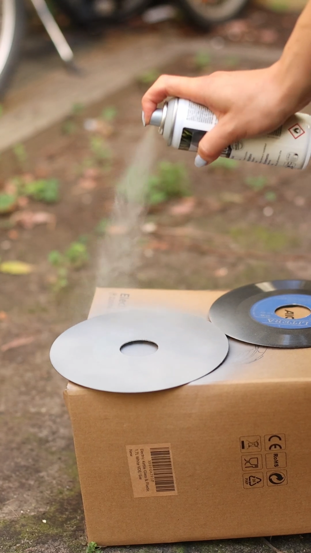 Eine Person verwendet die Upcycling-Idee „Badlampen“ von edding SHOP, um Vinyl-Schallplatten zu verwandeln und erzielt präzise Sprühlack-Ergebnisse. Zwei Schallplatten liegen auf einem Karton – eine bleibt schwarz, während die andere teilweise mit Effektspray Beton grau beschichtet ist. Der Hintergrund aus Gras und verstreuten Blättern verleiht dem kreativen Unterfangen eine natürliche Note.