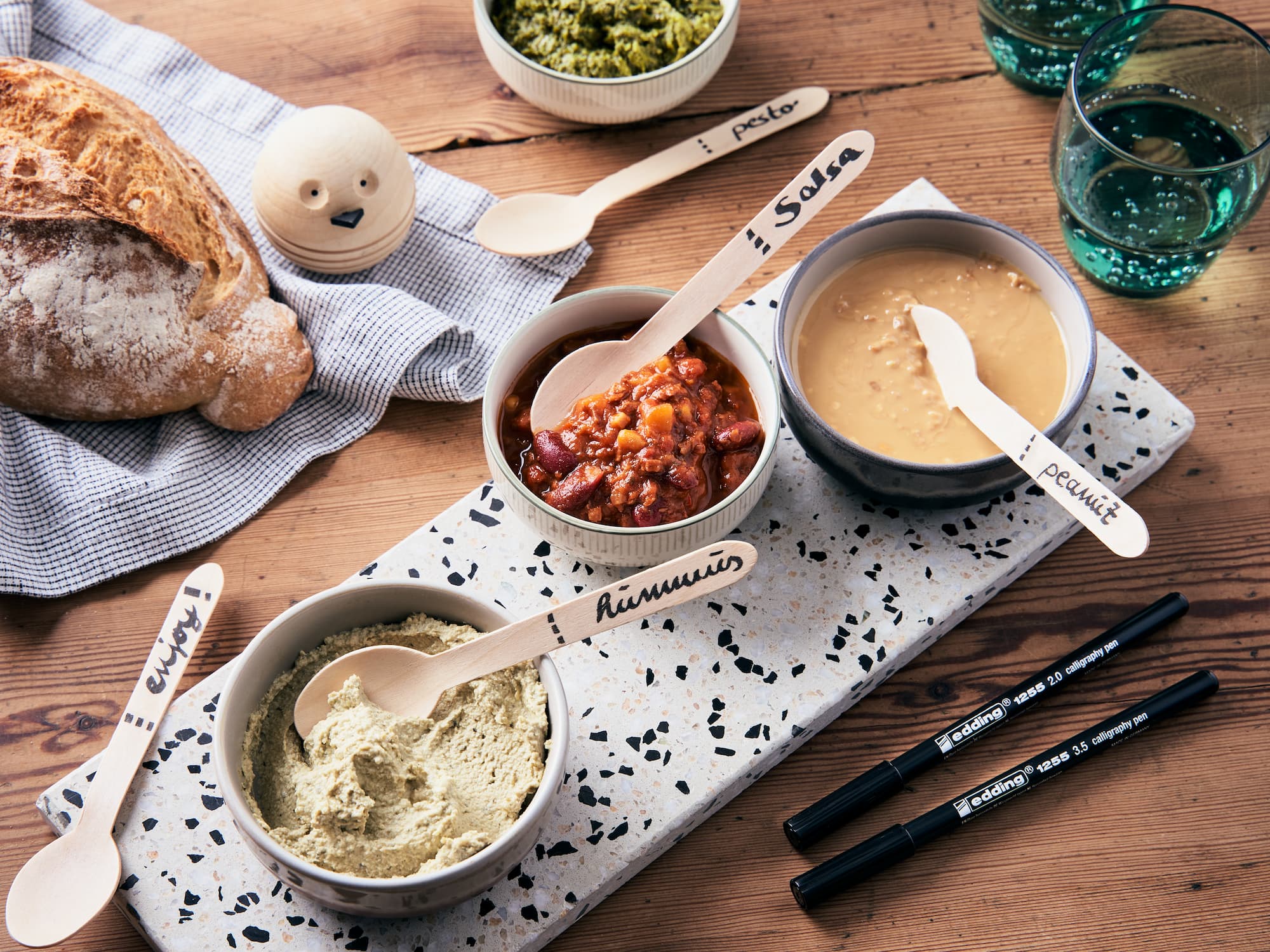 Auf einem Holzbrett stehen drei Schüsseln mit Pesto, Salsa und Erdnussdip, jeweils mit einem beschrifteten Holzlöffel. Auf dem Tisch stehen ein Laib Brot und Gläser mit Wasser, daneben ein edding 1255 Kalligrafiestift 3er-Set und einige schwarze Marker. Neben dem Brot liegt ein vogelförmiges Objekt.