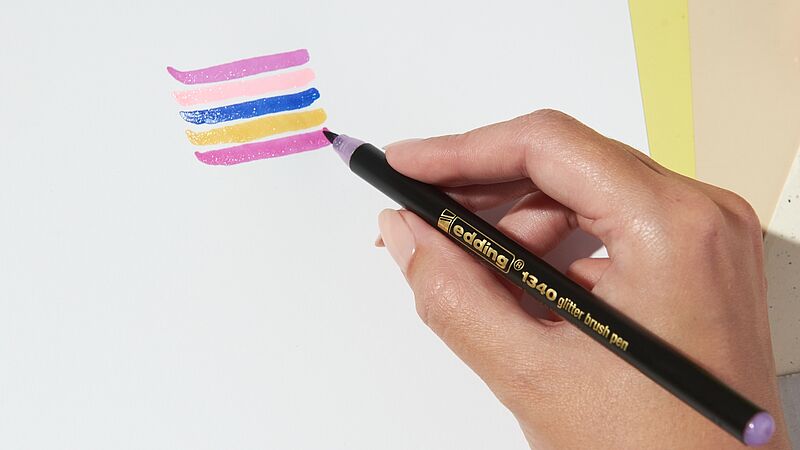 Eine Packung edding 1340 glitzer Pinselstift 10er-Set, ideal für Handlettering, präsentiert eine Vielzahl lebendiger Farben mit kunstvollen Kritzeleien auf der Vorderseite, die den glitzernden Tinteneffekt demonstrieren.