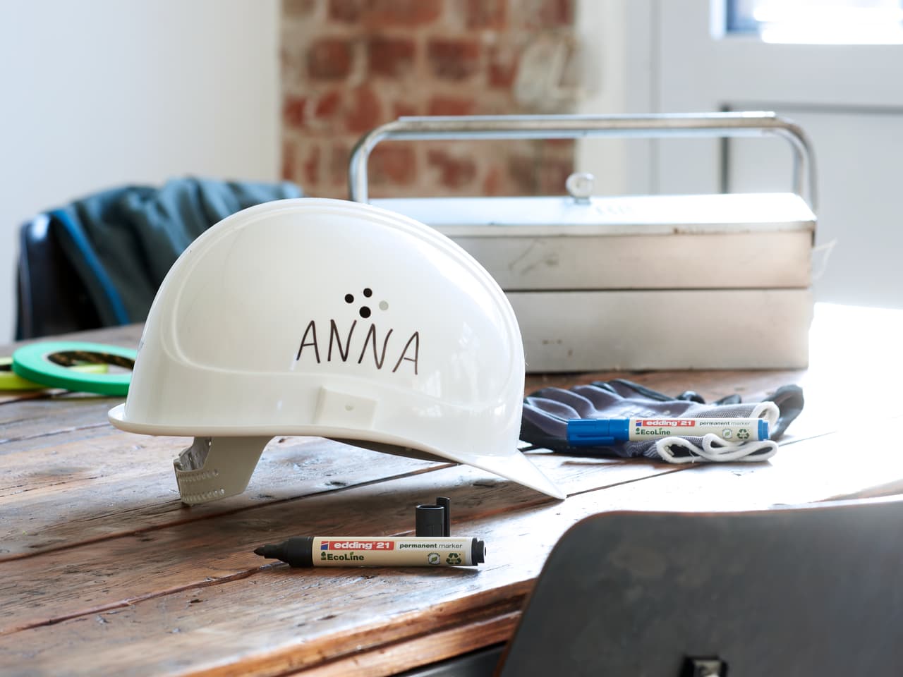 Ein weißer Schutzhelm mit dem Namen „Anna“, aufgedruckt mit einem edding 21 EcoLine Permanentmarker, platziert auf einem Holztisch neben einem Werkzeugkasten und Bauwerkzeugen in einem gut beleuchteten Behälter