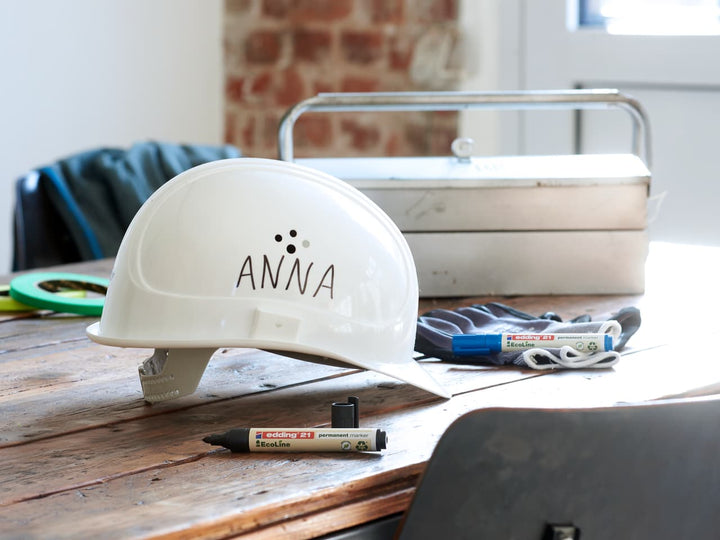 Ein weißer Schutzhelm, auf dem der Name „anna“ mit einem Edding 21 EcoLine Permanentmarker aufgedruckt ist, steht auf einem Holztisch inmitten verschiedener Werkzeuge und eines Werkzeugkastens aus Metall und lässt auf eine professionelle oder DIY-Projektumgebung schließen.