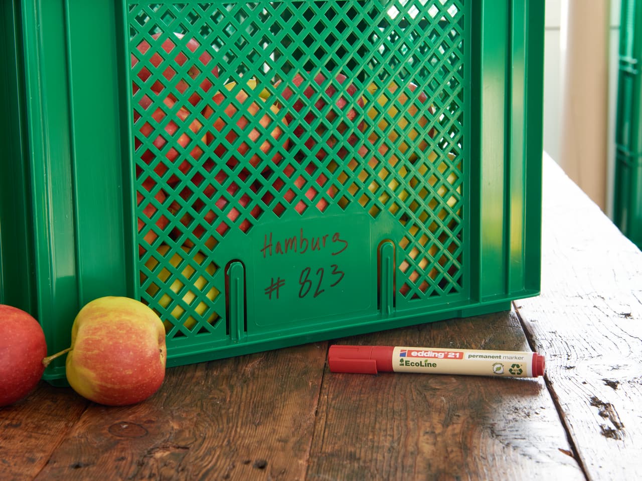 Grüne Plastikkiste mit der Aufschrift „hamburg #623“ daneben ein edding 21 EcoLine Permanentmarker und Äpfel auf einem Holztisch.
