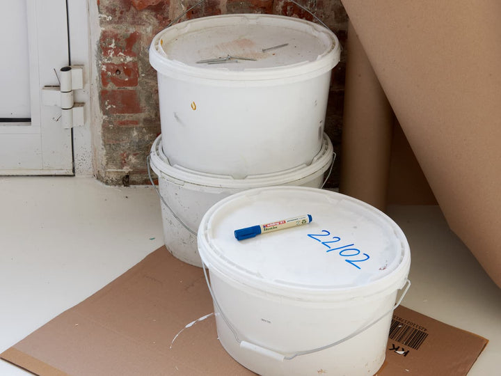 Drei gestapelte weiße Plastikeimer mit Deckel in einer überfüllten Ecke, wobei ein Eimer auf der Seite auf einem Stück Pappe steht und auf diesem Eimer ein edding 21 EcoLine Permanentmarker 4er-Set steht.