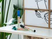 Ein aufgeräumter Arbeitsplatz mit dem edding 21 EcoLine Permanentmarker 4er-Set, hergestellt aus recyceltem Material, auf einem weißen Regal liegend, vor einer gerahmten abstrakten Strichzeichnung, mit einer grünen Pflanze als Akzent