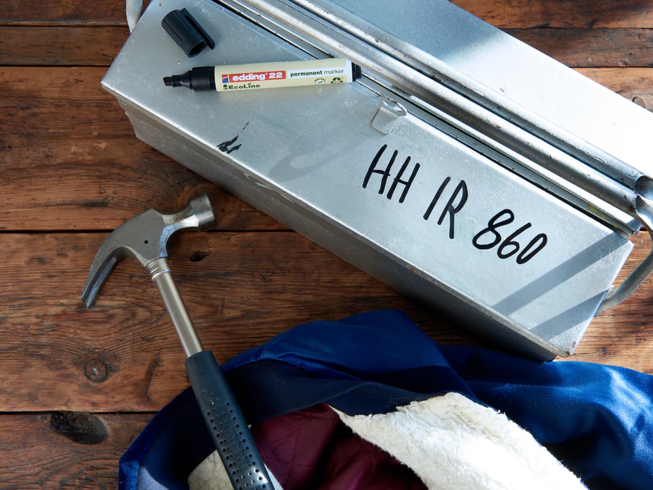 Ein Werkzeugkasten aus Metall mit der Aufschrift „hh ir 860“ neben einem Hammer und blauem Edding-Stoff auf einer Holzoberfläche.