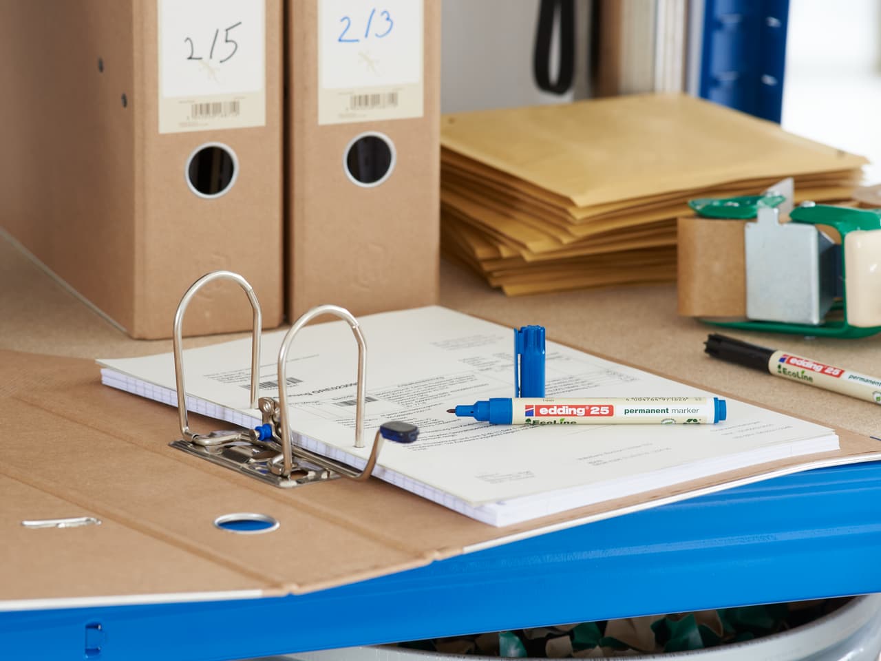 Ein offener Ringordner mit Dokumenten auf einem Schreibtisch, umgeben von Büromaterialien, darunter edding 25 EcoLine Permanentmarker, Stiften und Aufbewahrungsboxen aus Pappe.