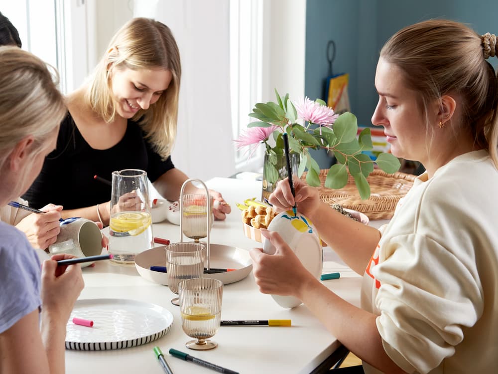 Drei Frauen bemalen kreativ Keramik an einem mit Snacks und Blumen geschmückten Tisch und genießen einen kreativen und geselligen Nachmittag mit edding 4200 Porzellanpinselstiften.