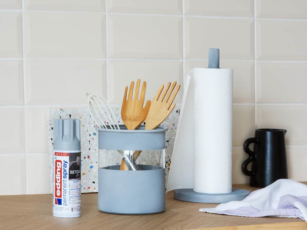 Eine aufgeräumte Küchenarbeitsplatte mit einem Satz Holzutensilien in einem Halter, einer Küchenpapierrolle auf einem Ständer und Reinigungsmitteln einschließlich einer Dose edding 5200 Permanentspray Effekt Acryllack.