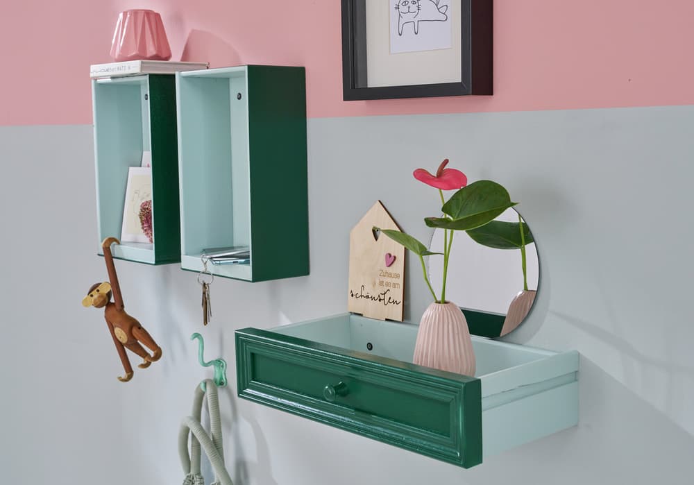 Ein stilvoller Wandorganizer mit einer Schublade aus edding 5200 Permanentspray Acryllack Neon & kräftige Farben, grünen Schränken und einem Akzentregal aus Holz. Eine rosa Vase, ein kleiner Spiegel und Dekorationsgegenstände, darunter eine Affenfigur, sind ordentlich angeordnet.