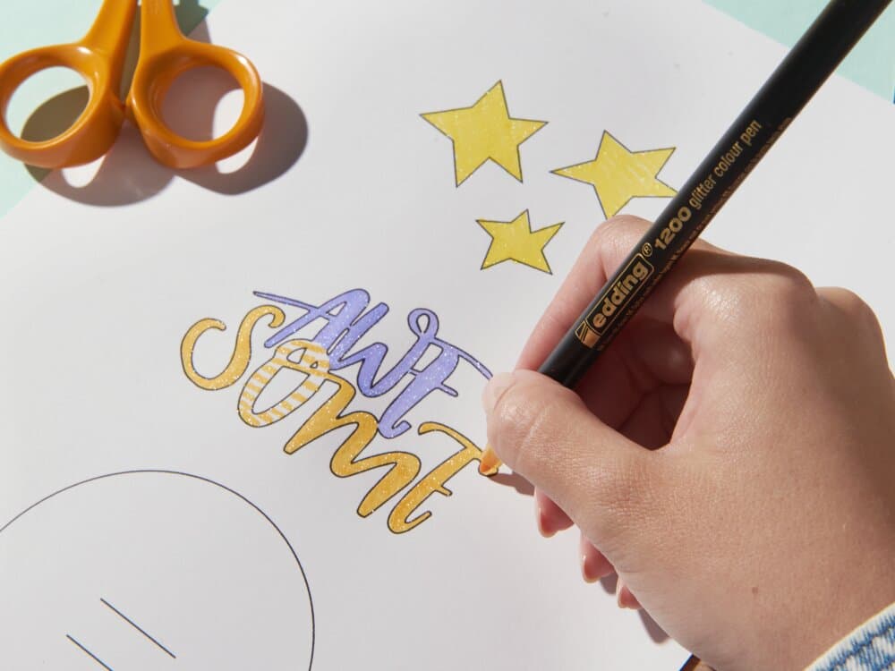 Die Hand einer Person hält einen edding 1200 Glitzer Fasermaler und schreibt das Wort „awesome“ in bunten Buchstaben auf ein Blatt Papier mit gezeichneten Sternen und einer Schere in der Nähe.