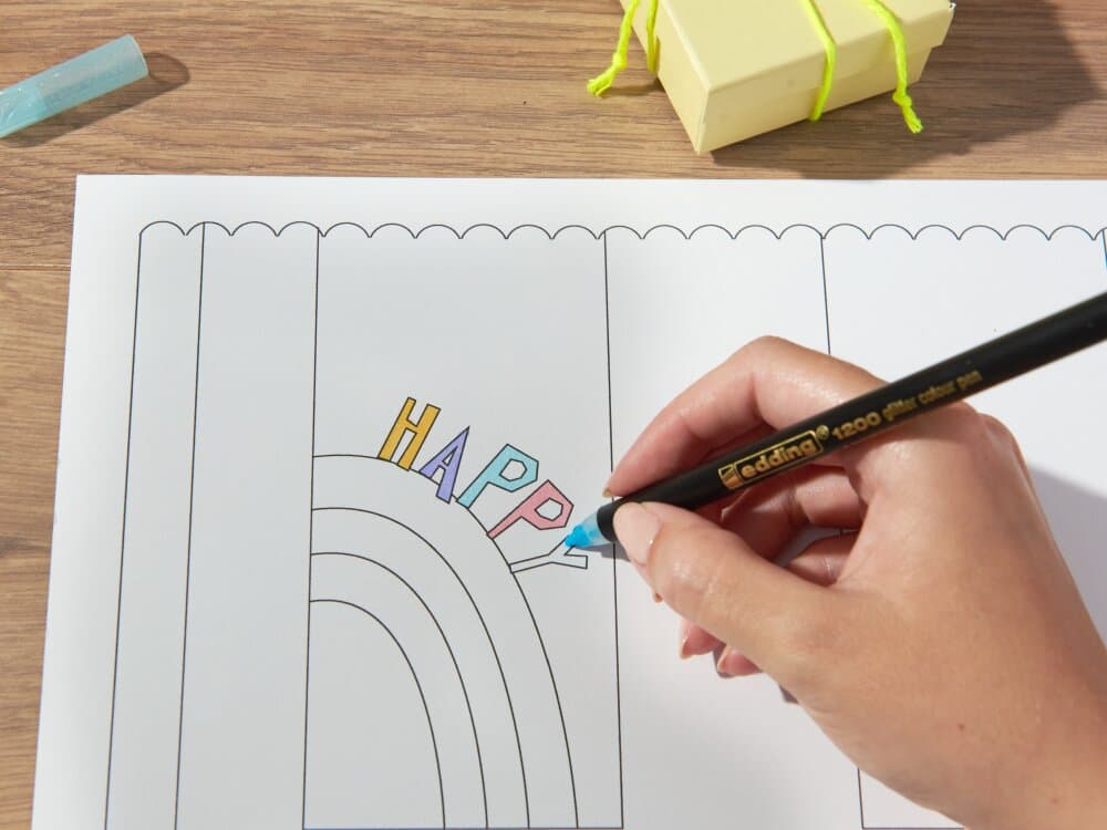 Die Hand einer Person malt das Wort „happy“ auf einer Malseite mit Regenbogenmotiv mit einem schwarzen Glitzer-Fasermaler edding 1200 aus. Auf dem Tisch liegen außerdem ein Radiergummi und eine gelbe Origami-Figur.