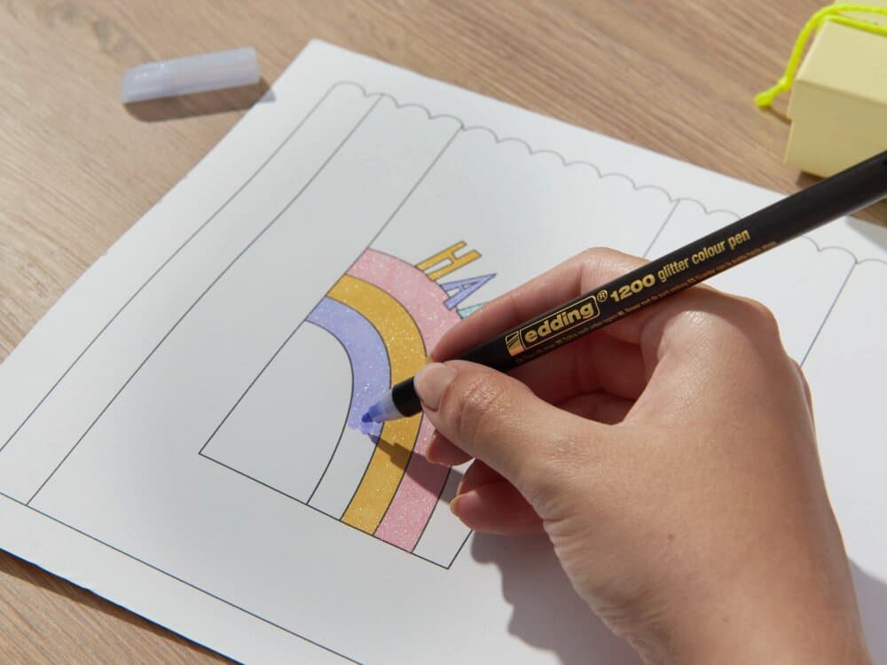 Die Hand einer Person malt mit einem schwarzen Buntstift einen Regenbogen auf ein Malblatt. Auf einem Holztisch stehen ein Edding 1200 Glitzer Fasermaler und ein Anspitzer in der Nähe.