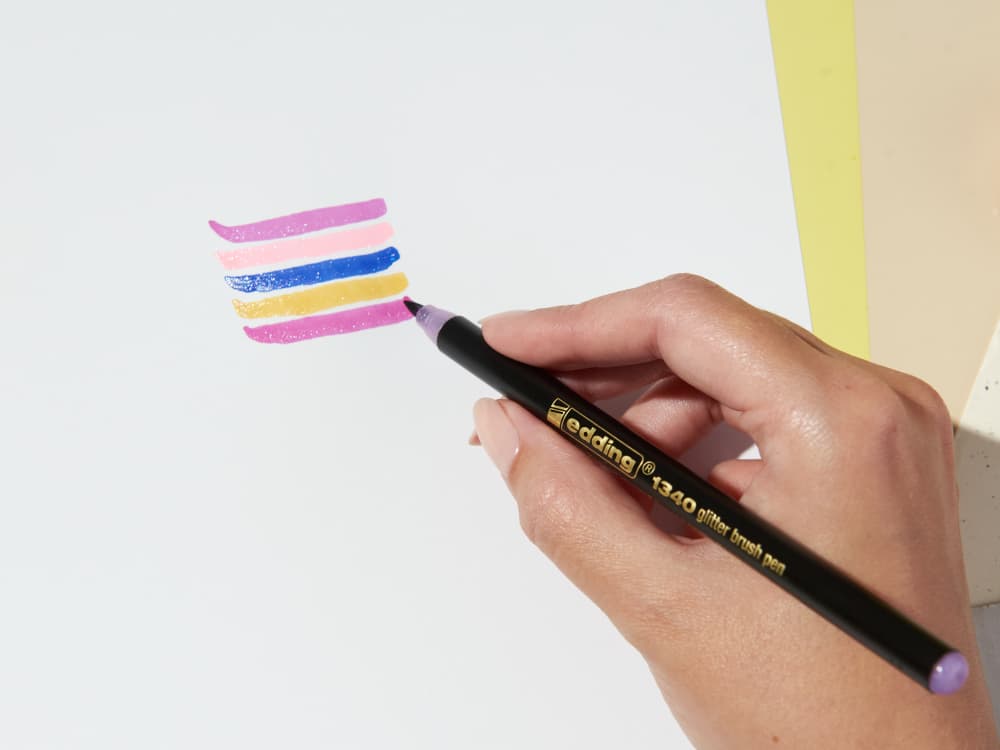 Eine Person hält einen edding 1340 Glitzer-Pinselstift in der Hand und trägt damit rosa, weiße, gelbe und blaue horizontale Streifen auf eine helle Oberfläche auf.
