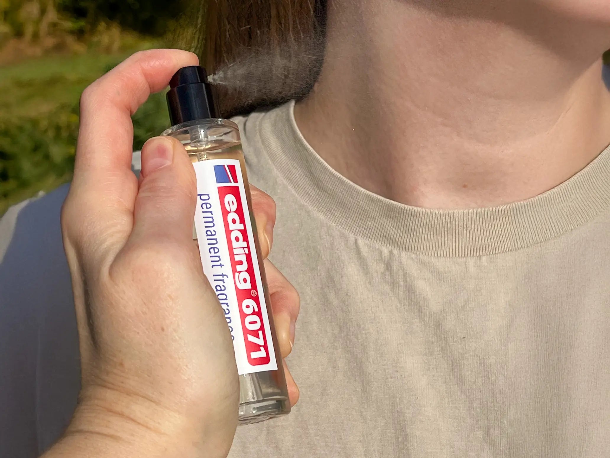 Eine Person, die im Freien ein beigefarbenes Hemd trägt, sprüht sich bei Tageslicht den edding 6071 permanent fragrance by edding seitlich in den Nacken.