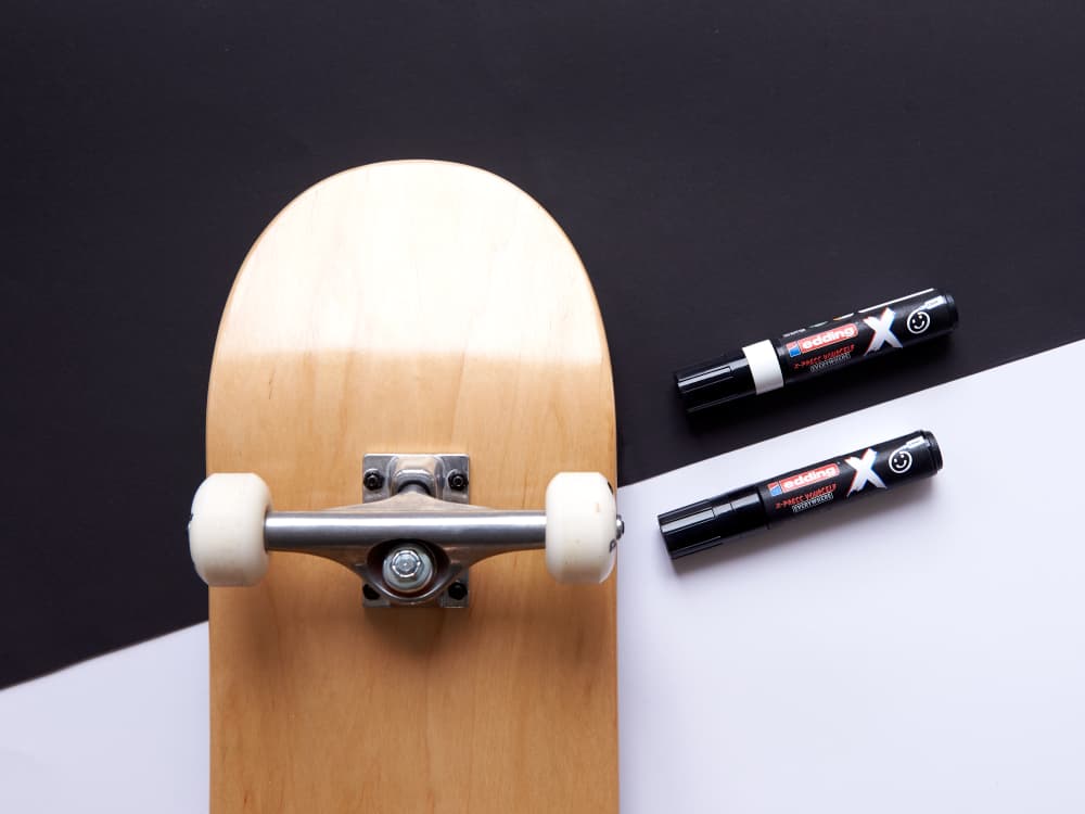 Auf einem schwarz-weißen Hintergrund steht ein Skateboard mit weißen Rädern und einer natürlichen Holzoberfläche. Neben dem Skateboard befinden sich zwei edding 6900 X Multimarker Black & White Color-Stifte von edding – einer liegt und der andere steht aufrecht, beide mit langlebiger Tinte.