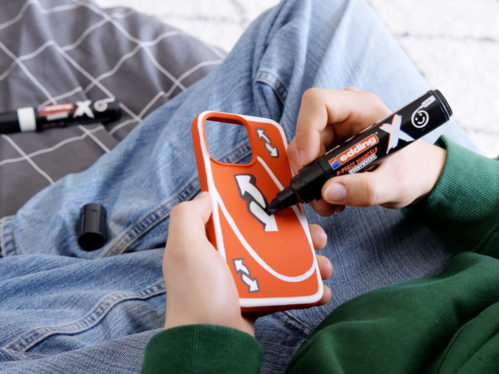 Eine Person hält eine orangefarbene Handyhülle mit weißen Pfeilen und zeichnet mit einem edding 6900 X Multimarker Schwarz aus dem Black & White Color 2er-Set darauf. In Blue Jeans und grünem Top sitzt die Person, die im Hintergrund eine gemusterte Decke trägt, die für eine gemütliche Note sorgt. Daneben liegen eine Markerkappe und ein weiterer vielseitiger Keilspitze-Marker von edding.