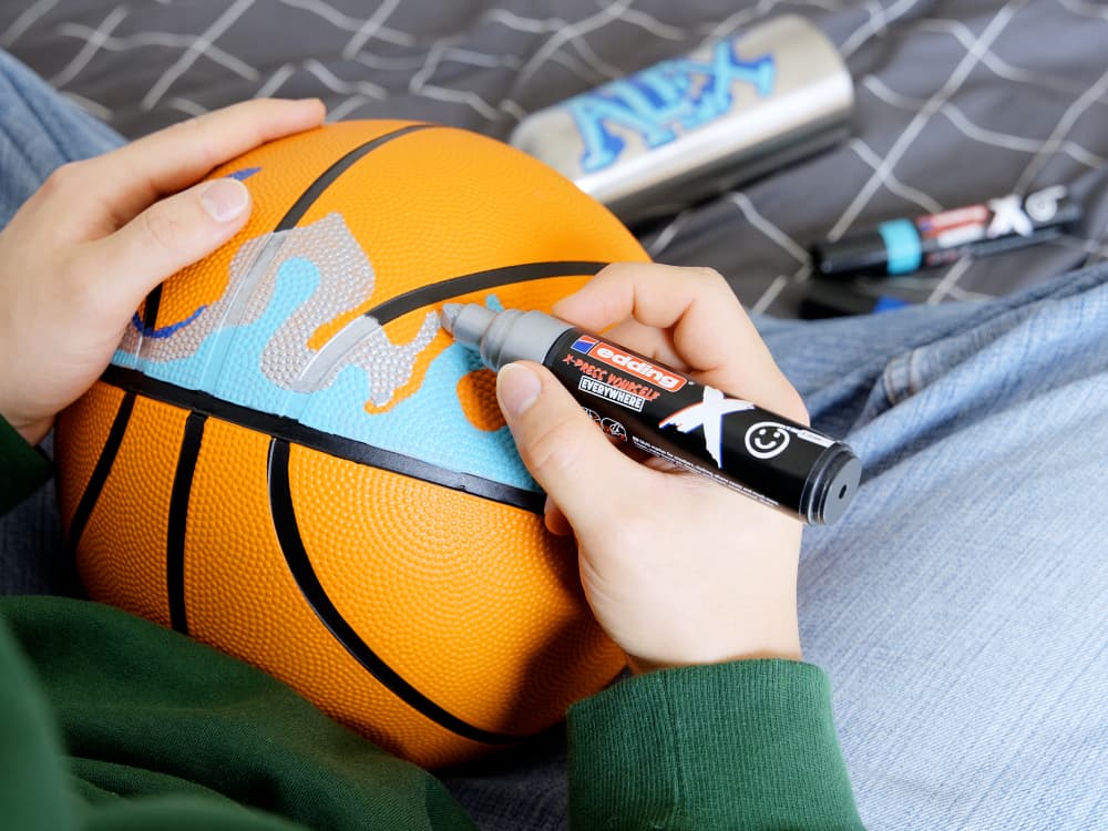 Eine Person personalisiert einen orangefarbenen Basketball mit einem edding 6900 X Multimarker Ocean Color 3er-Set. Der Ball wird mit einem blau-grauen Design verziert. Es sieht so aus, als ob die Person auf einem Bett mit einer schwarz-weißen Decke mit geometrischem Muster sitzt, umgeben von weiteren Markern mit langlebiger Tinte für anhaltende Kreativität.